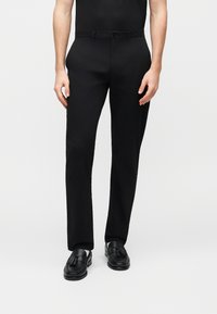 Μαύρο, ραμμένο παντελόνι με λεία υφή και ίσια γραμμή ποδιού. Διαθέτει κούμπωμα με κουμπί και θηλιές για ζώνη. Φοριέται με μαύρα loafers.