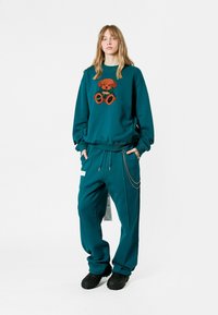 Sudadera color verde azulado con bordado de oso, combinada con pantalones holgados en el mismo color que cuentan con cordón y detalle de cadena, luciendo con zapatos negros texturizados.