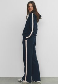 Woman wearing navy blue tracksuit with white side stripes and white sneakers, standing sideways against a plain wall, hands in pockets.