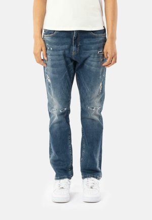 Jean en denim bleu avec des détails usés, présentant des motifs de déchirure et une coupe décontractée, associés à des baskets blanches.