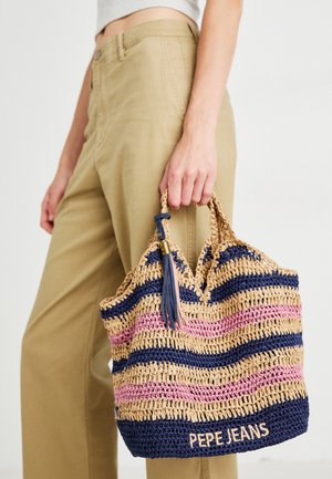 Main tenant un sac fourre-tout tricoté avec des rayures bleues, roses et beiges et la marque "PEPE JEANS", porté avec un pantalon beige et un haut blanc.