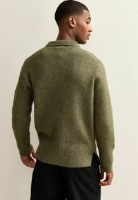 Suéter de punto verde oliva con cuello de chal, tela texturizada y puños acanalados, visto desde la parte de atrás, llevando sobre pantalones negros.