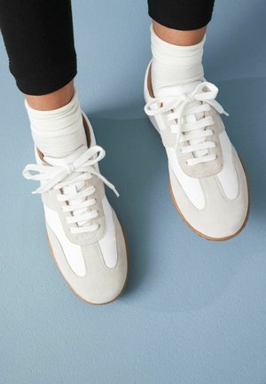 White and gray athletic shoes feature a suede and leather upper, white laces, and a brown rubber sole. Worn with white crew socks.