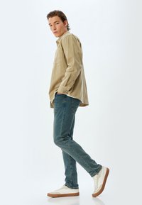 Camicia beige a maniche lunghe con tasca, indossata sopra jeans di denim blu. Sneakers bianche con dettagli color crema. Il modello è in piedi di lato su uno sfondo bianco.