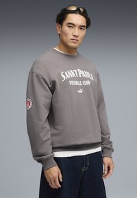 Sudadera Puma, algodón gris, cuello redondo, con "SANKT PAULI FUSSBALL CLUB" en blanco, puños acanalados, emblema en la manga, ajuste relajado.