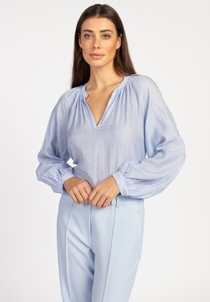 Himmelblaue Bluse mit langen Puffärmeln und tiefem V-Ausschnitt, aus einem strukturierten Stoff, kombiniert mit himmelblauen, maßgeschneiderten Hosen.