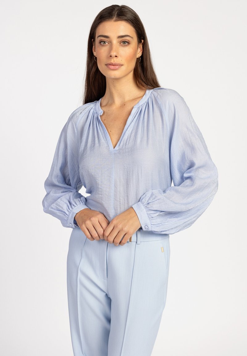 Himmelblaue Bluse mit langen Puffärmeln und tiefem V-Ausschnitt, aus einem strukturierten Stoff, kombiniert mit himmelblauen, maßgeschneiderten Hosen.
