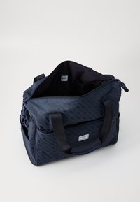 Marineblå duffelbag med et geometrisk mønster, dobbelthåndtag, lynlåslukning og et logo-patch. Rummeligt interiør med en mørk foring.