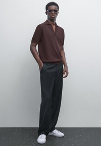 Brun kortærmet polo skjorte med en tekstureret design, parret med sorte brede bukser og hvide sneakers, mod en lys baggrund.