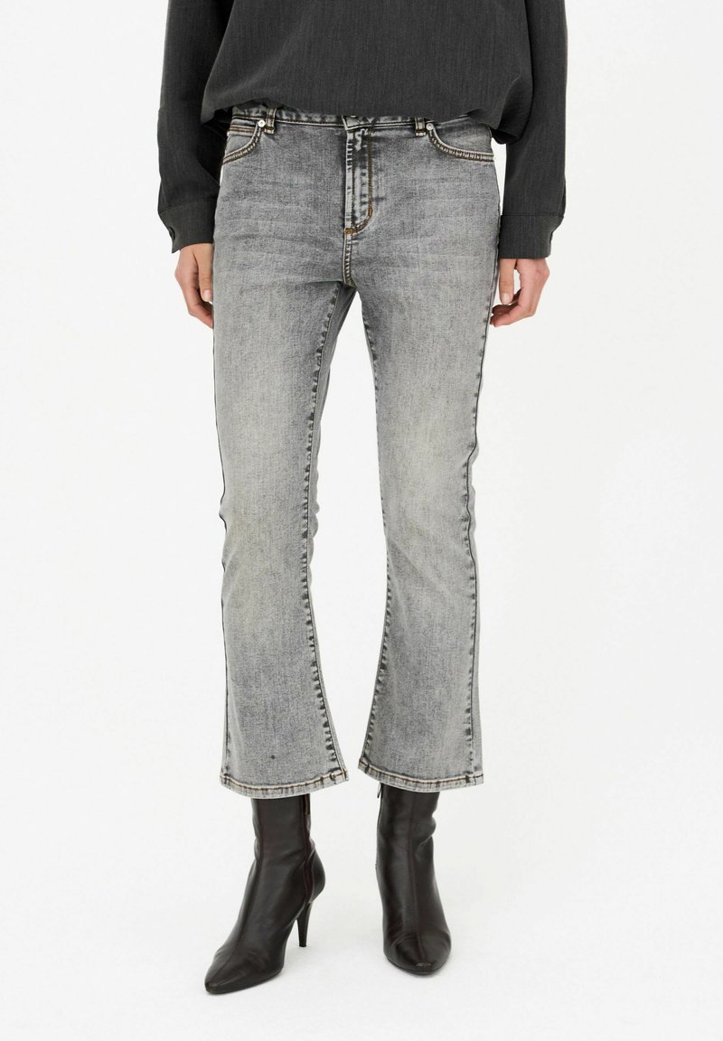 Personne portant un jean bootcut gris délavé, un top noir à manches longues et des bottines noires à talons aiguilles et bout pointu.
