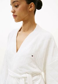 Witte doorgestikte badjas met een sjaalkraag, ceintuurafsluiting en een klein geborduurd logo op de borst. Zachte textuur en losse pasvorm.