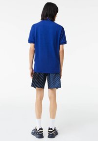 Personne aux cheveux noirs mi-longs portant un t-shirt bleu à manches courtes, un short bleu et noir à motifs, des chaussettes blanches et des baskets noires, debout.