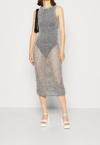 Šaty ke kolenům bez rukávů vyrobené z černé a bílé pletené síťoviny s transparentním designem. Doplněné o bílé platformové sandály a černou tašku.