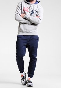 Felpa grigia con logo rosso e blu, pantaloni tapered blu navy e sneakers grigie con accenti neri. Calzini bianchi visibili sopra le scarpe.
