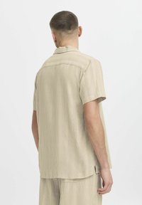 Camisa de manga corta a rayas en color beige con cuello. Textura suave con un ajuste relajado, presenta un dobladillo recto y aberturas en los costados.