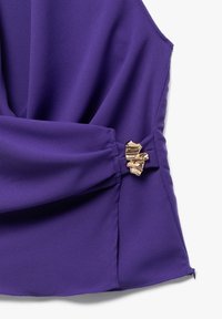 Top viola senza maniche con tessuto drappeggiato, caratterizzato da un accento decorativo dorato sul lato sinistro e un orlo ordinato lungo il bordo inferiore.