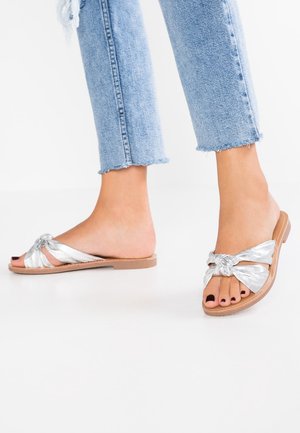 Sandales plates en métal argenté avec un design noué, semelle plate et bout ouvert. Portées avec un jean léger bleu déchiré.