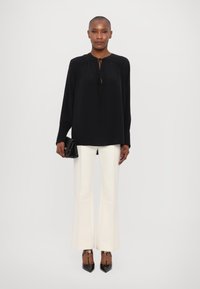 Blusa nera a maniche lunghe con dettaglio al collo a fiocco, abbinata a pantaloni bianchi svasati. Modella che tiene in mano una piccola pochette nera, indossando tacchi a punta neri.