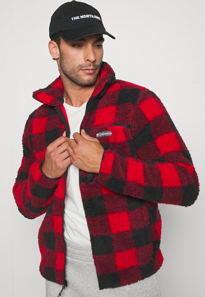 Veste en polaire à carreaux rouges et noirs avec fermeture éclair, col haut et détail de logo. Portée sur une chemise blanche, sur fond uni.