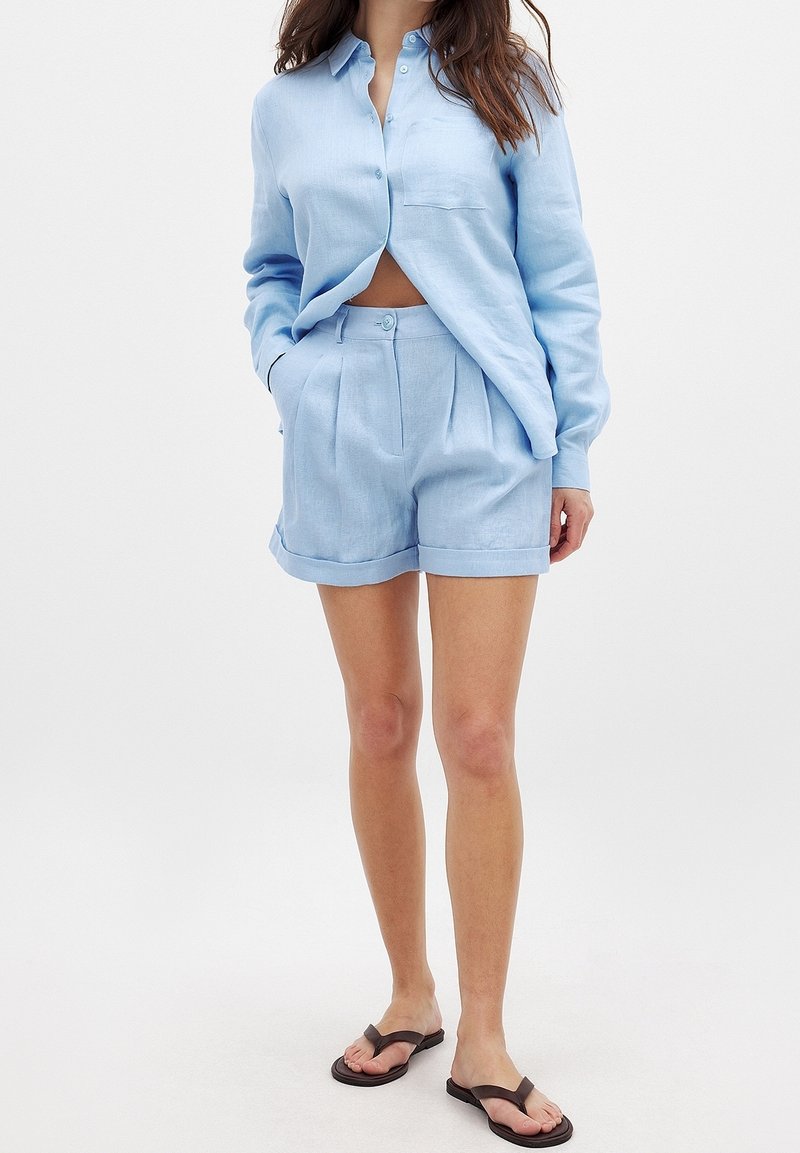 Chemise en lin bleu clair avec boutons et poche, associée à un short plissé assorti. Coupe confortable, texture lisse et ourlet retroussé.