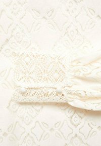 Bluse i blonde i off-white med indviklede blomster mønstre og tekstureret detaljer. Sammensunkne ærmer med knapdetaljer forbedrer designet.