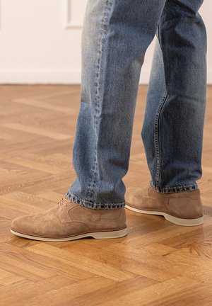 Persoon die beige suède nette schoenen en blauw spijkerbroek draagt, staand op een houten parketvloer.