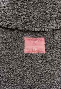 Šedá, huňatá látka s texturovaným povrchem. Na látce je přišitá růžová štítková látka s bílým textem a dekorativní grafikou.