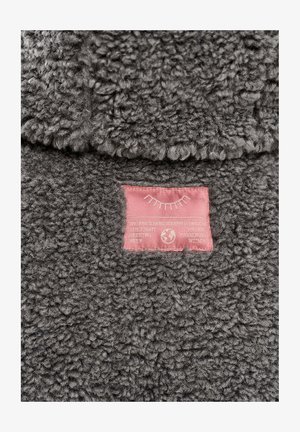 Szary, puszysty materiał o teksturowanej powierzchni. Na nim przyszyta jest różowa etykieta z białym napisem oraz dekoracyjną grafiką.