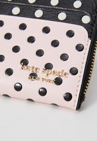 Portefeuille noir et rose à pois, fabriqué en cuir texturé, avec des détails en métal doré et un logo de marque embossé sur un fond rose.