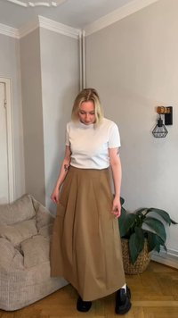 Balta īss piedurkņu T-krekls kopā ar brūnu krokotu midi svārku, valkāts ar melniem apaviem; iekštelpu vide ar augiem un mēbelēm fonā.