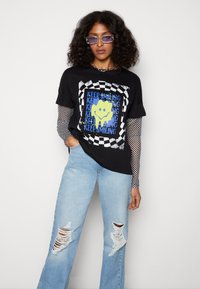 T-shirt gráfica preta com um design de rosto sorridente amarelo e texto azul "CONTINUE A SORRIR". Usada sobre uma blusa de mangas longas xadrez. Jeans claros desgastados.