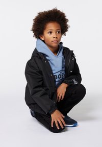 Veste noire imperméable avec capuche, superposée à un sweat-shirt bleu clair arborant un logo, accompagnée d'un pantalon noir et de chaussures navy.