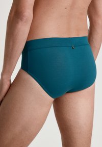 Turkooise herenslip gemaakt van zacht materiaal, met een naadloos ontwerp en een elastische tailleband met een klein goudkleurig detail aan de achterkant.