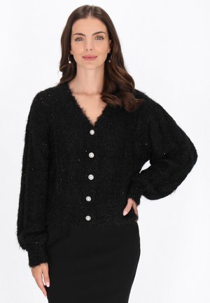 Vrouw met lang bruin haar draagt een zwarte pluisachtige cardigan met parelknopen, staand met één hand op haar heup tegen een witte achtergrond.
