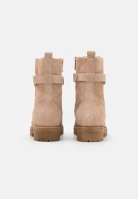 Bottines en daim beige avec une finition texturée, présentant des double sangles, des accents dorés et une semelle en caoutchouc empilée.