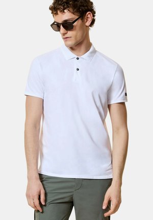 Giovane uomo con occhiali da sole neri, indossa una polo bianca a maniche corte con bottoni e pantaloni verde oliva, in piedi con postura rilassata.