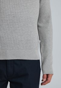 Pulover tricotat texturat de culoare gri deschis, cu manșete și tiv ondulat, purtat cu pantaloni de culoare albastru închis, arătând brațul și mâna dreaptă relaxate.