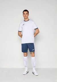 Modelo masculino con cabello corto que lleva una camiseta de fútbol blanca y azul marino, pantalones cortos azul marino, calcetas blancas hasta la rodilla y botas blancas, posando contra un fondo liso.