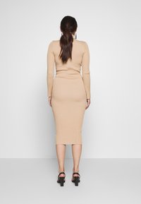Abito bodycon lavorato a maglia in beige con maniche lunghe, texture a coste e lunghezza a metà polpaccio. Indossato con tacchi neri a cinturini.