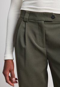 Olijfgroene broek met plooien, voorzien van een knoopsluiting en lussen voor een riem. Gemaakt van een gestructureerde stof, samen met een getailleerde witte langemouwtop.