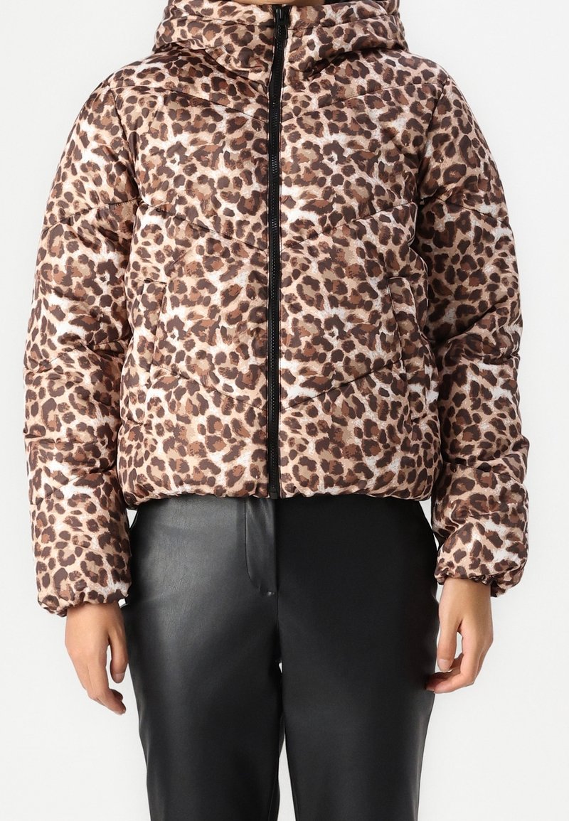 Kurtka puchowa w panterkę z kapturem, w odcieniach brązu i kremu, zapinana na zamek, z elastycznymi mankietami. Noszona z czarnymi skórzanymi spodniami.