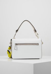 Borsa rettangolare bianca con manico superiore, tasca con zip frontale e sciarpa a motivo giallo-nero legata sul lato, posizionata su un piedistallo bianco.