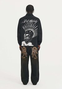 Chaqueta negra con gráfico de calavera blanca y rayos de sol en la espalda; pantalones negros de corte holgado con bordado de dragón naranja en los lados.