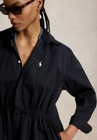 Kvinne med flettefrisyre som har på seg solbriller, en svart kjole med krage og en hvit logo, et blått perlearmbånd og en hvit skjelløredobb.