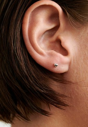 Közeli kép egy bal fülről, rövid barna hajjal és egy kis ezüst szív alakú fülbevalóval a fülcimpán.