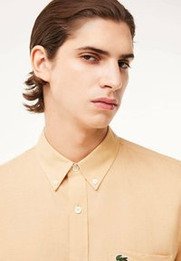 Camicia button-up color beige chiaro realizzata in tessuto leggero, con colletto a punta e un piccolo logo ricamato sulla tasca del petto.
