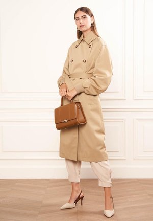 Manteau beige à double boutonnage et ceinture à la taille, associé à un pantalon clair et des escarpins beiges à bout pointu. Sac à main marron en accent.