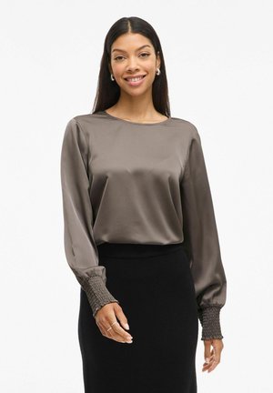 Femme aux longs cheveux foncés souriante, portant un chemisier en satin taupe à manches longues avec poignets smockés et une jupe noire taille haute.