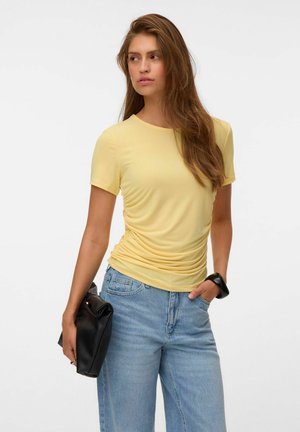 Donna con capelli castani lunghi che indossa una maglietta gialla arricciata a maniche corte e jeans celesti, tiene una pochette nera e indossa un braccialetto nero.