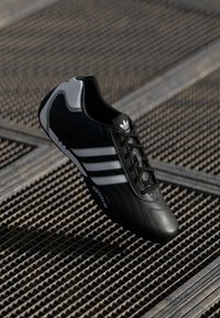 Black Adidas sneaker with white stripes and logo, displayed above a metal grated surface.
